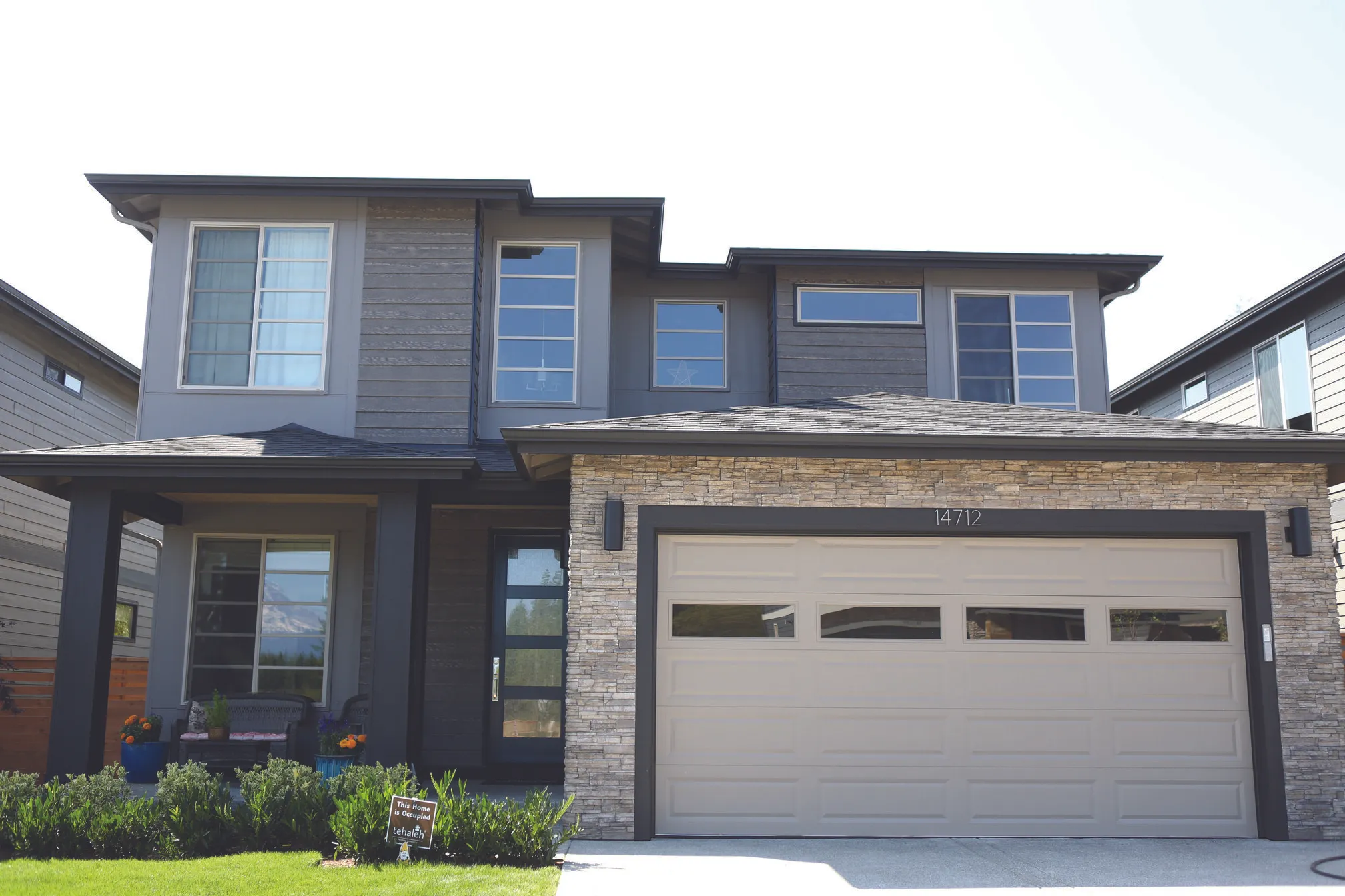 Modern two-story house with stone accents, a beige garage door, and a small front porch with plants and seating.